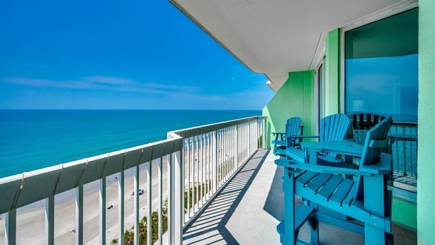 A balcony with a view of the beach and ocean, featuring blue Adirondack chairs and a white awning. - Oceanfront Modern Home w Lazy River and Pools (North Myrtle Beach)