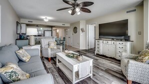A living room with a sofa, armchair, coffee table, TV, and a ceiling fan. - Oceanfront Modern Home w Lazy River and Pools (North Myrtle Beach)
