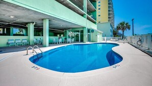 A swimming pool with a curved edge, a covered parking area, and a building with a green facade and multiple floors. - Oceanfront Modern Home w Lazy River and Pools (North Myrtle Beach)