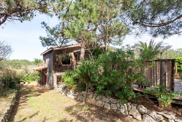 A rustic wooden house with a stone wall, surrounded by dense greenery and trees.