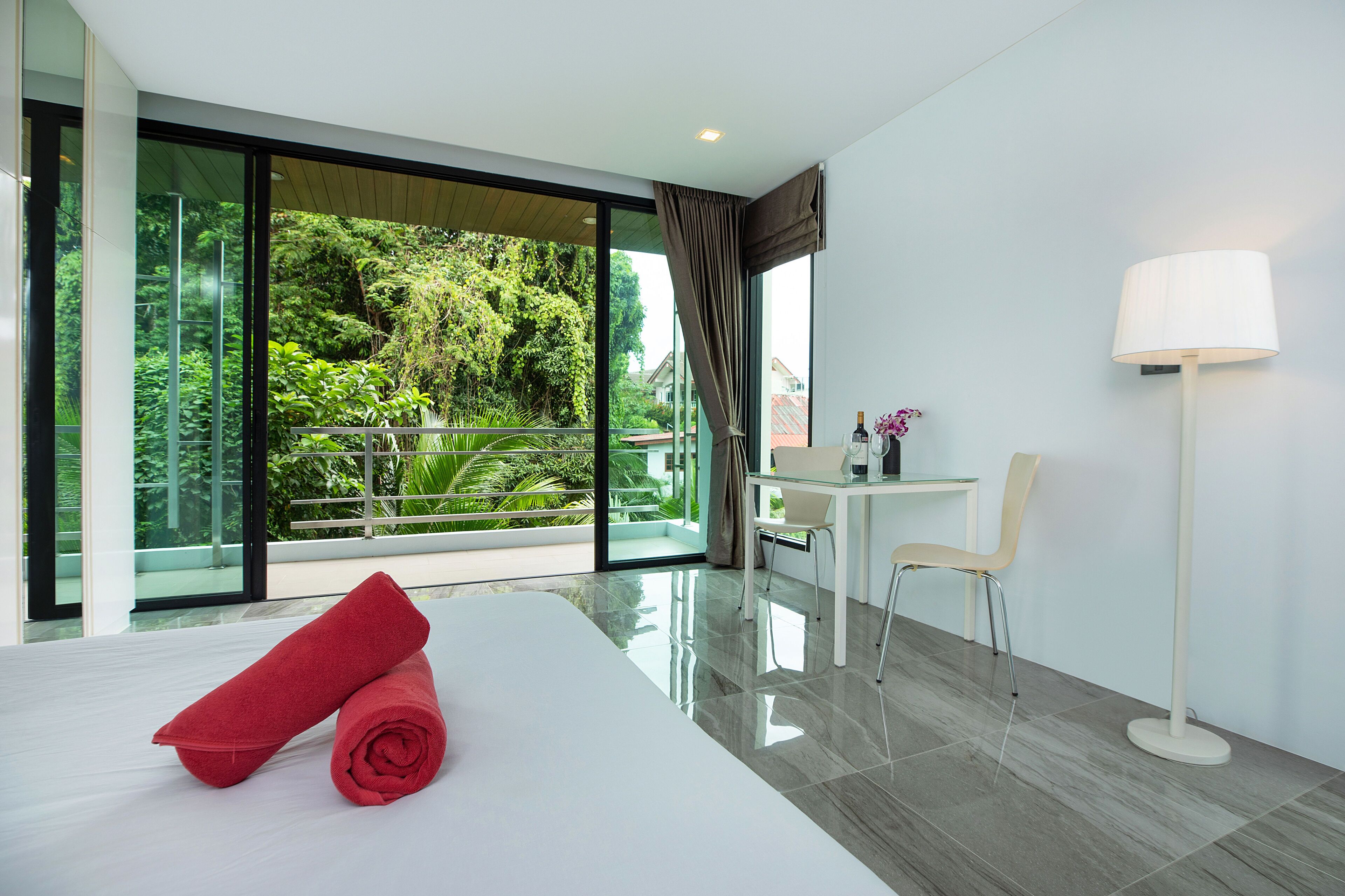A modern hotel room with a large bed, a desk, a chair, and a view of lush greenery through large windows.