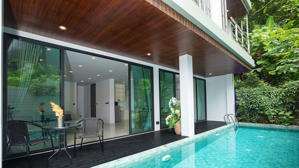 A modern outdoor pool area with a wooden deck, glass doors leading to a dining area, and a view of lush greenery. - Kata Pool Villa UTK5 for Beach Lovers  (Karon)