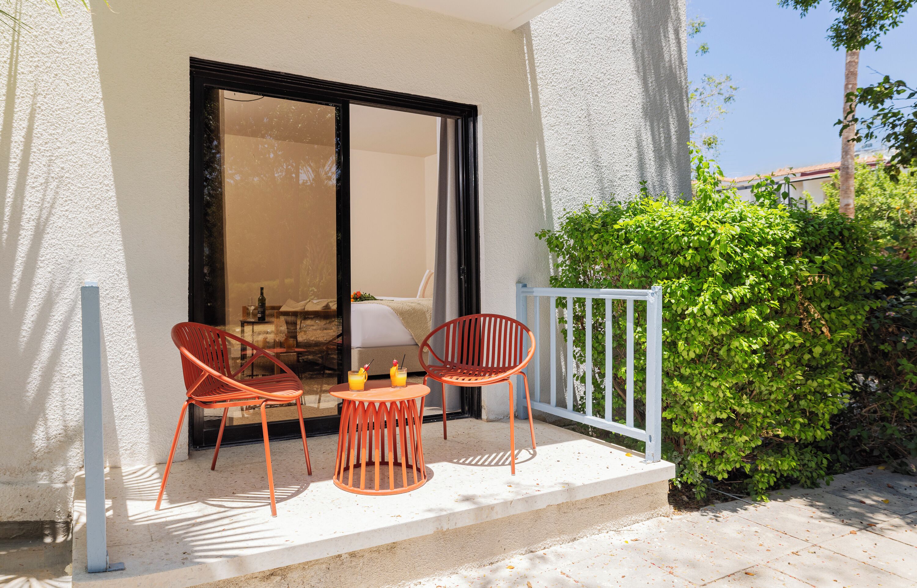 A balcony with two chairs and a small table, a sliding glass door, and a green hedge.