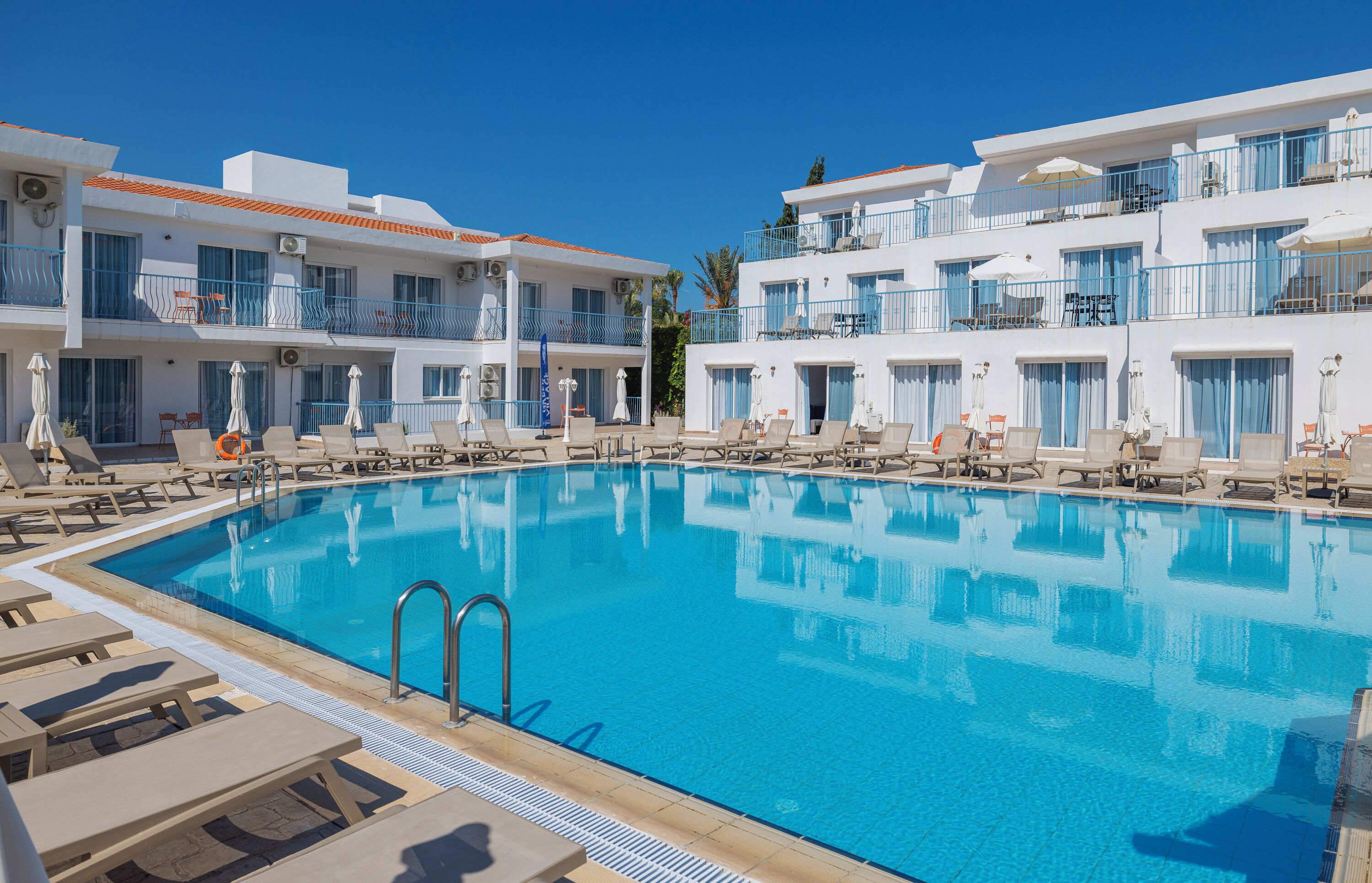 Une piscine entourée de bâtiments blancs dotés de balcons, avec des chaises longues et des parasols à proximité.