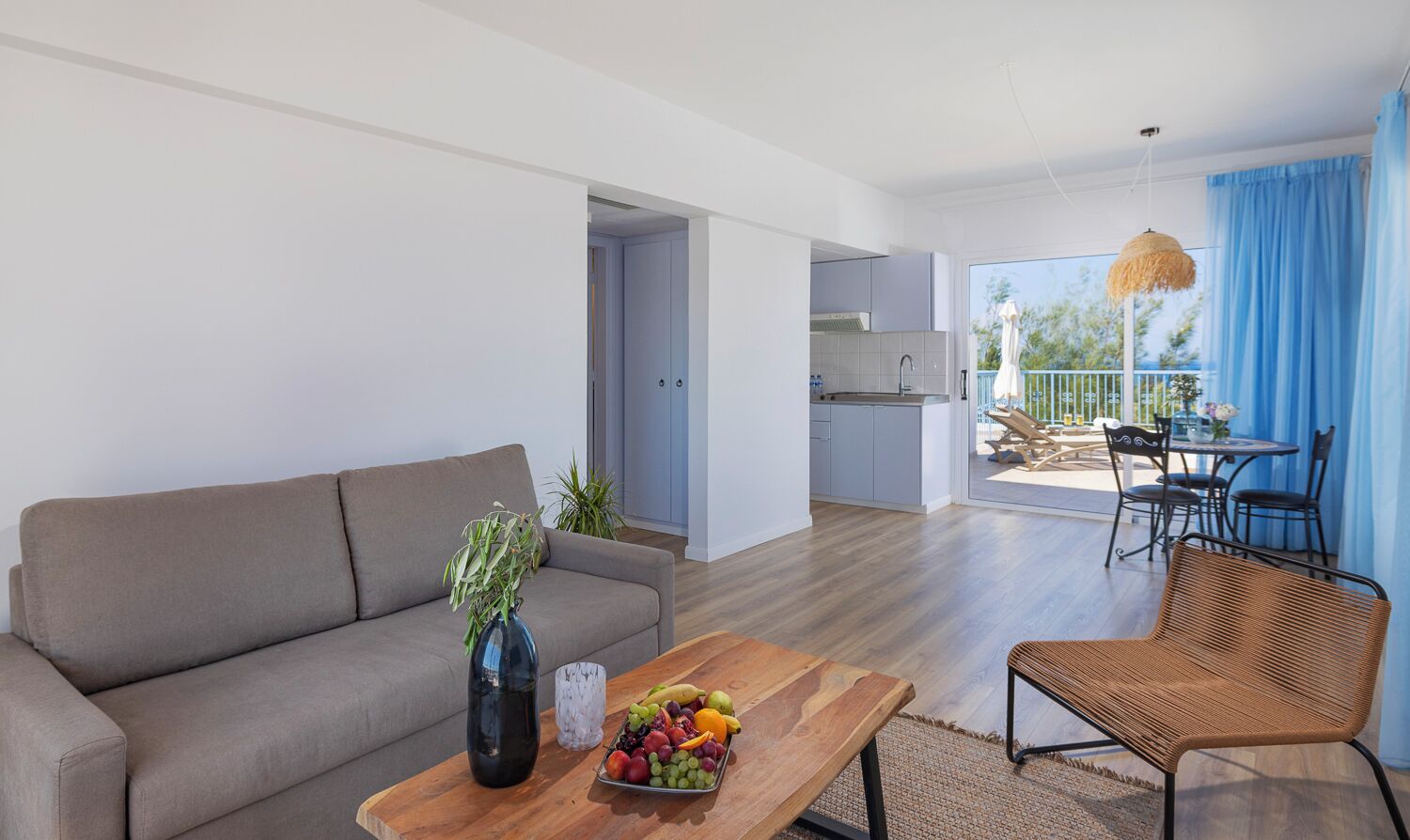 Una sala moderna con un sofá, una mesa de centro con fruta, una silla de mimbre y vistas al balcón y la cocina.