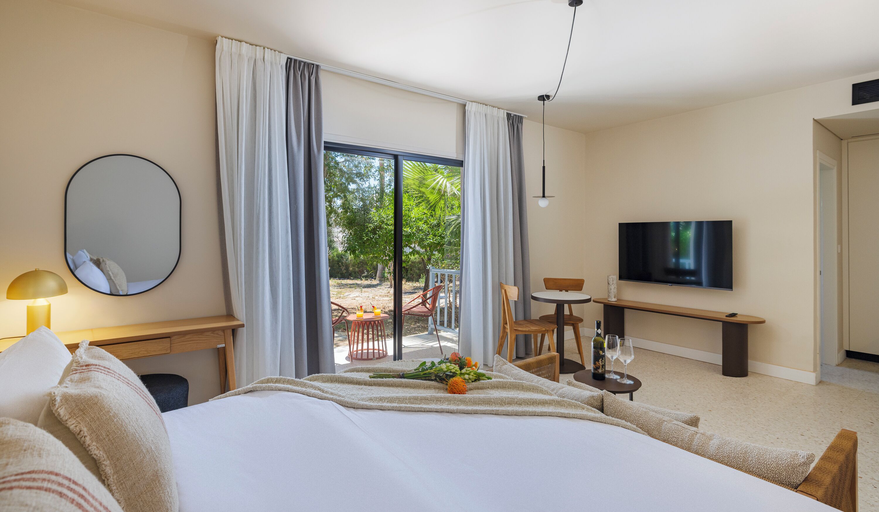 Habitación de hotel con cama, escritorio, silla, televisión y balcón con vistas a árboles.