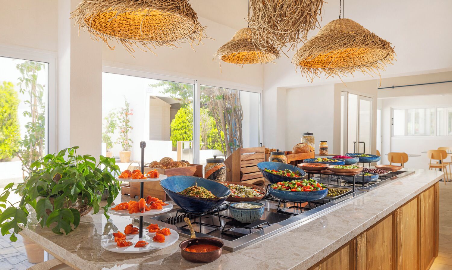 A buffet setup with various dishes, a large plant, and wicker ceiling lights.