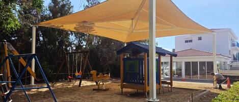Un parque infantil con columpios y una zona sombreada, rodeado de árboles y un edificio.