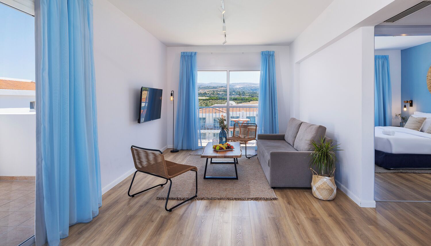 A modern living room with a sofa, a dining table, and a chair. There is a large window with curtains, a flat-screen TV, and a view of buildings outside.