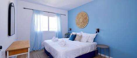 A bedroom with a bed, a bedside table, a small round table, a wall-mounted lamp, and a window with curtains.