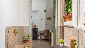 A narrow hallway with a stone sink, potted plants, a small table, and a door leading to a room with a table and chairs. - Apartment 'Giulia's Home - Borgo' with Private Terrace, Wi-Fi and Air Conditioning (Catania)