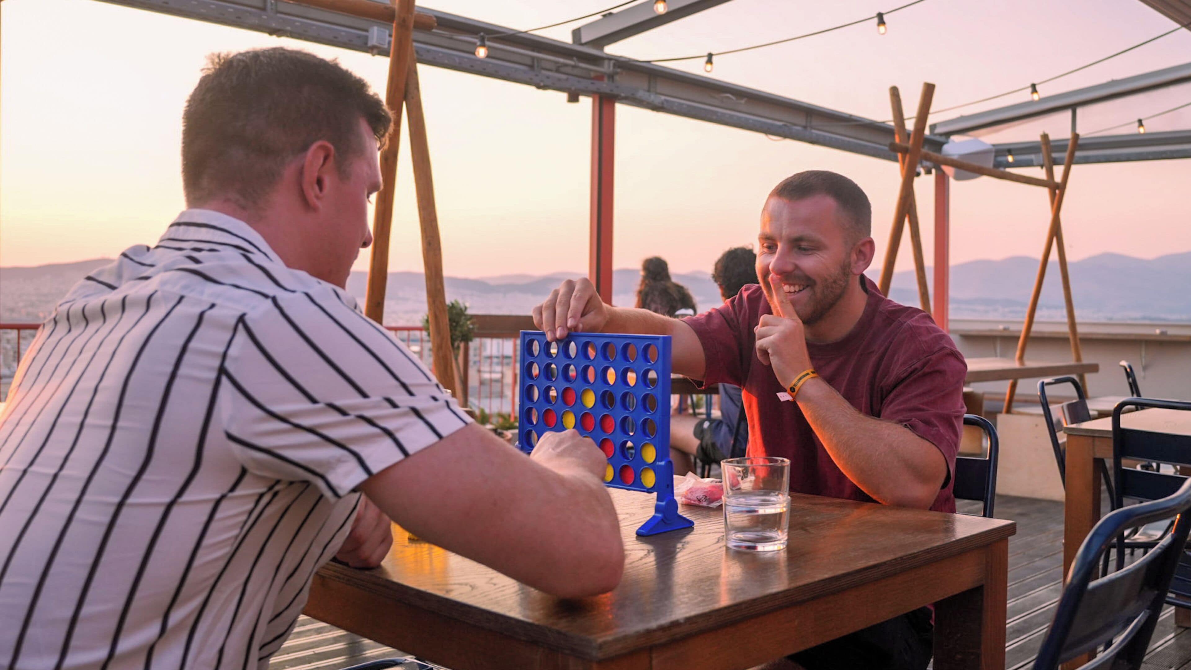 Two men playing a board game on an outdoor table with a view of mountains in the background.