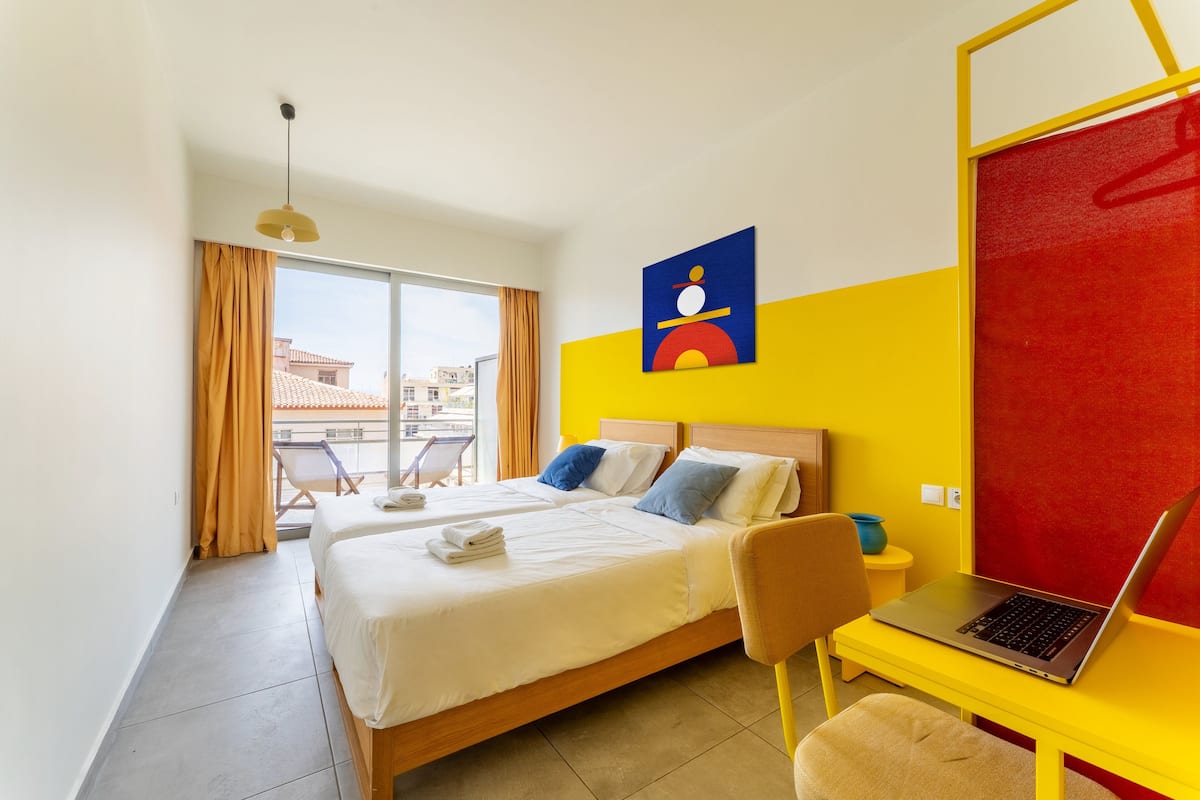 Une chambre d’hôtel avec deux lits, un bureau avec un ordinateur portable, une vue sur les bâtiments environnants et un mur de séparation rouge.