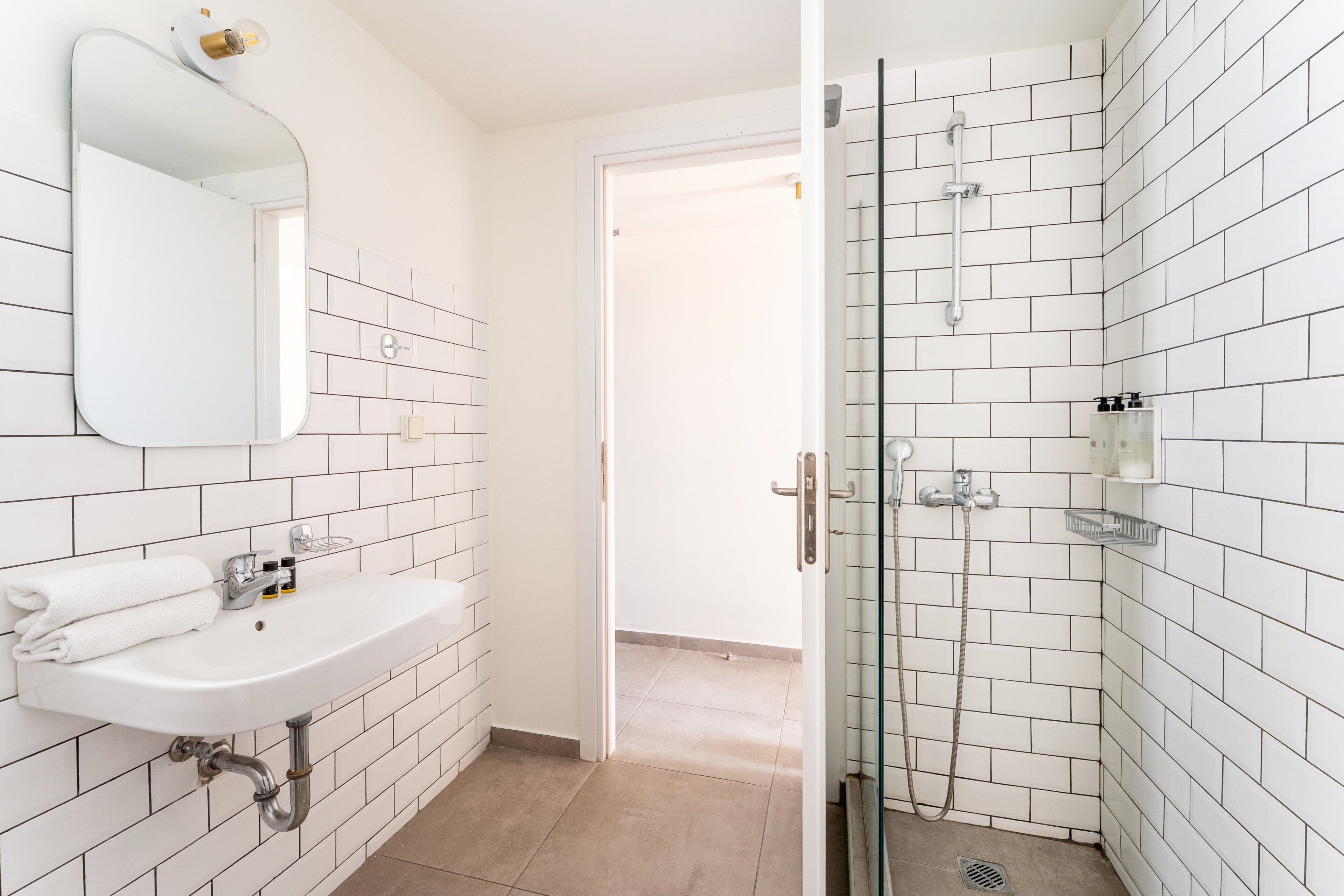 A bathroom with white tiled walls, a sink with a mirror, and a shower area.