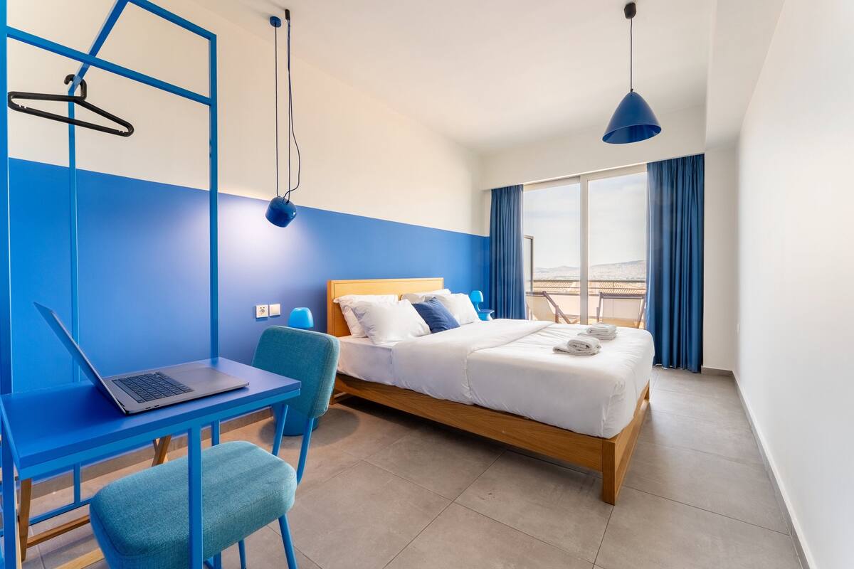 Une chambre d’hôtel moderne équipée d’un lit, d’un bureau avec un ordinateur portable, d’une chaise et offrant une vue sur l’extérieur.