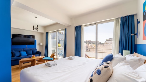 A hotel room with a large bed, a sofa, a TV, and a balcony with blue curtains. - YellowSquare Athens (Athens)