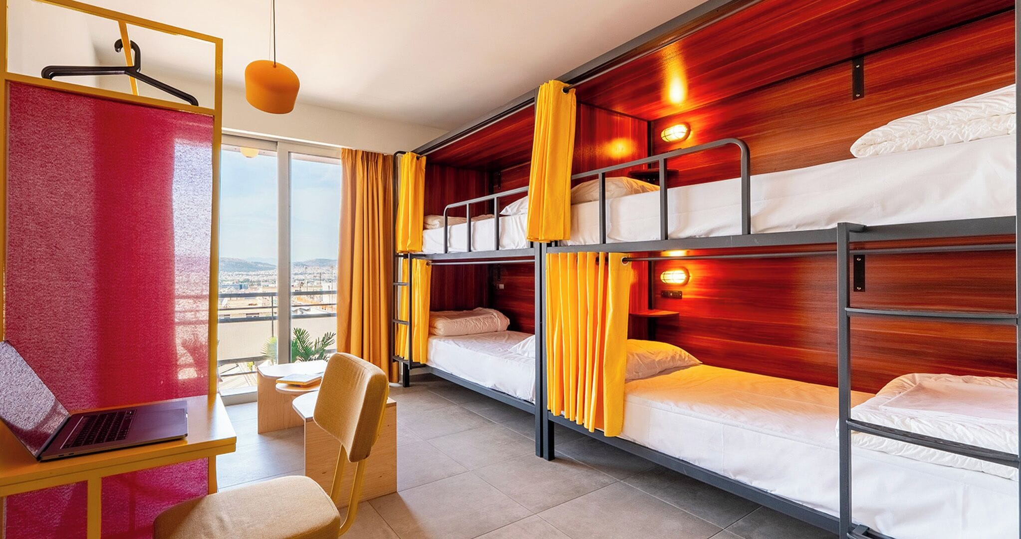 A hotel room with bunk beds, a desk with a laptop, a chair, and a view of the city.