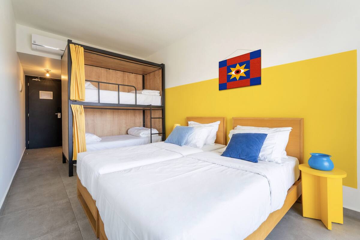 Une chambre d’hôtel avec un lit superposé, deux lits simples, une petite table de chevet jaune et un drapeau accroché au mur.