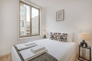 Une chambre d’hôtel avec un lit, deux lampes de chevet, une fenêtre donnant sur des immeubles et un tableau encadré au mur.