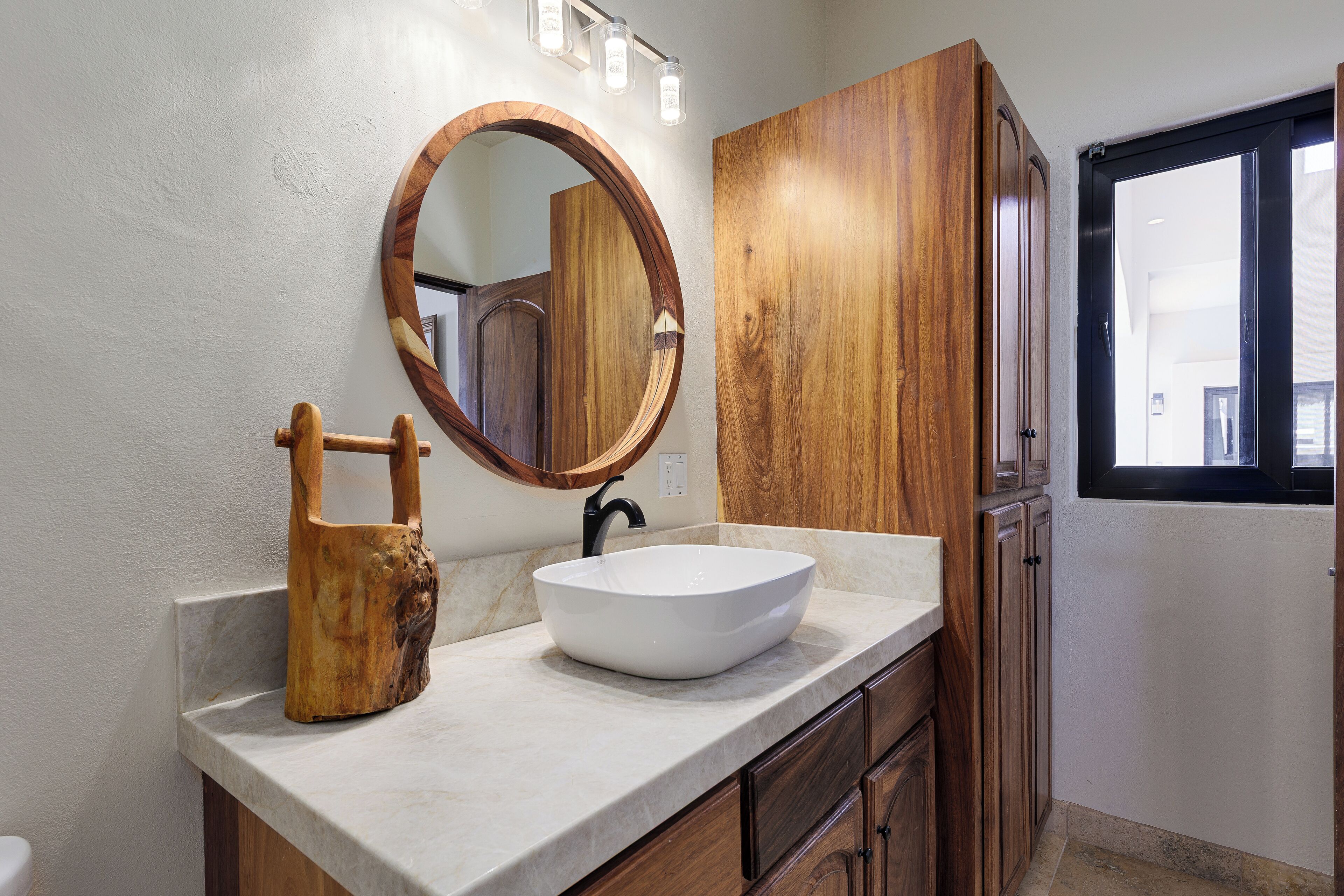 A bathroom with a white marble countertop, a wooden vanity, a round mirror, and a black faucet.