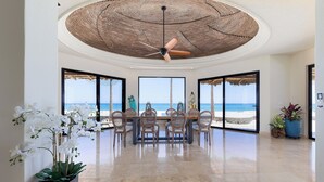 Una habitación amplia con una gran mesa de comedor, sillas y un ventilador de techo. Amplias ventanas ofrecen vistas a la playa.
