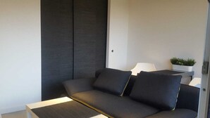Een moderne woonkamer met een donkergrijze bank, een klein bijzettafeltje met een lamp en een potplant op een plank.