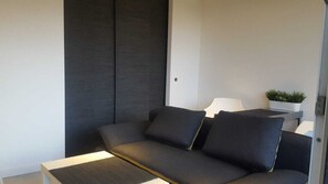 A modern living room with a dark-colored sofa, a small side table with a lamp, and a potted plant on a shelf.