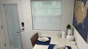 Een eethoek met een tafel, stoelen, een raam met luxaflex, een wandlamp en een decoratief object.