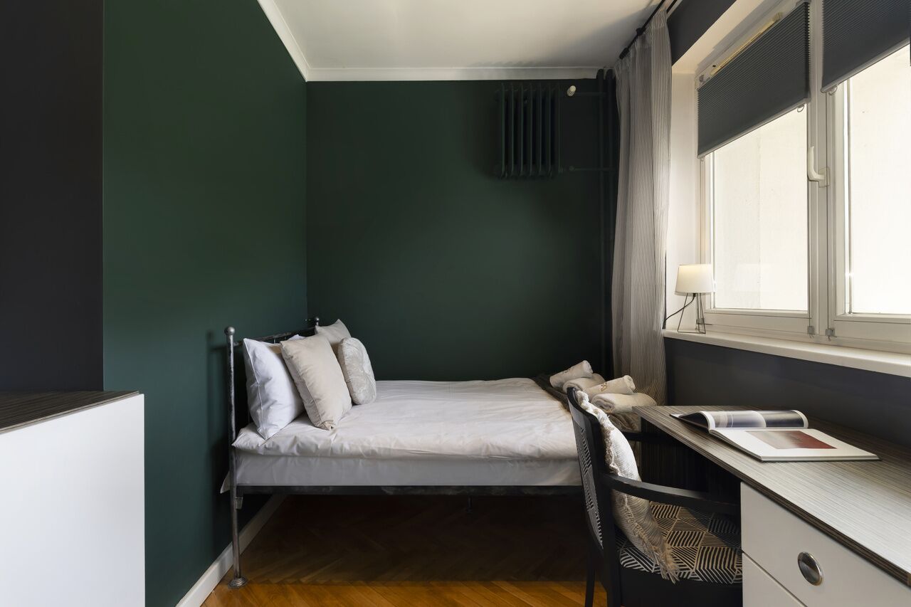 Une chambre à coucher de taille modeste, comprenant un lit simple, un bureau avec une chaise, une fenêtre avec des rideaux et un radiateur.