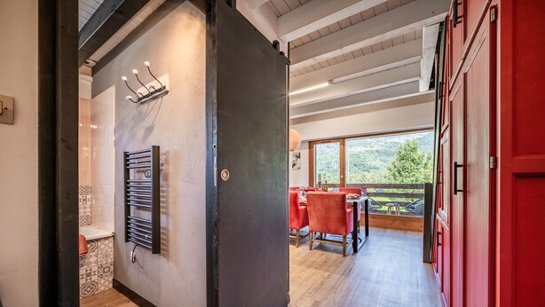 A modern interior with a wooden floor, a dining area with red chairs, and a view of greenery outside.