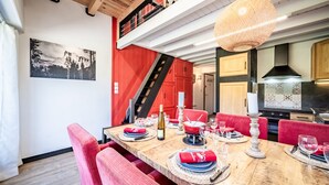 A dining area with a wooden table set for six, red upholstered chairs, a staircase, and a kitchen in the background.