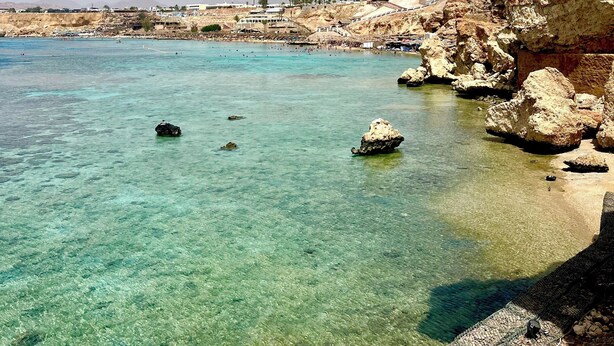 A coastal scene with clear turquoise waters, rocky cliffs, and a distant town. - Red Sea Breeze · Modern 1BR in Hadaba · Sharm El Sheikh (South Sinai Governorate)