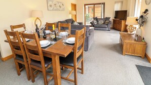 Un coin repas avec une table en bois dressée pour quatre personnes, un canapé gris, un téléviseur et une petite table d’appoint avec une lampe et des objets décoratifs.