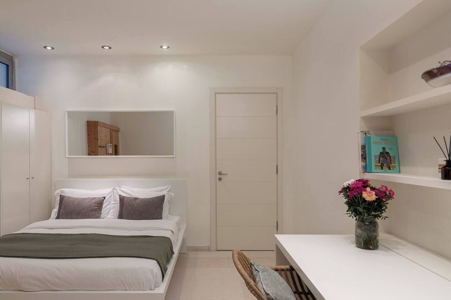 Un dormitorio con cama, mesita de noche, espejo, puerta y estante con un libro y un florero.
