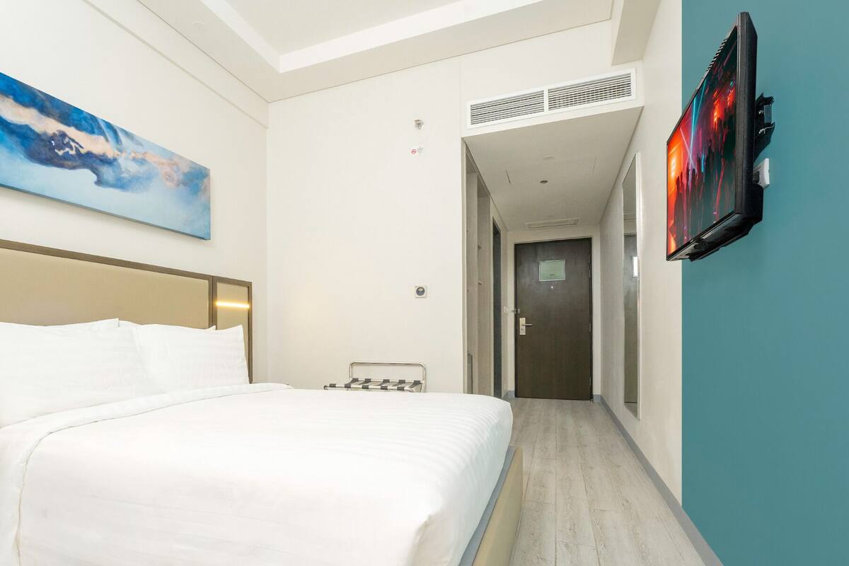 A hotel room with a bed, a painting, a television, and a hallway leading to another room.