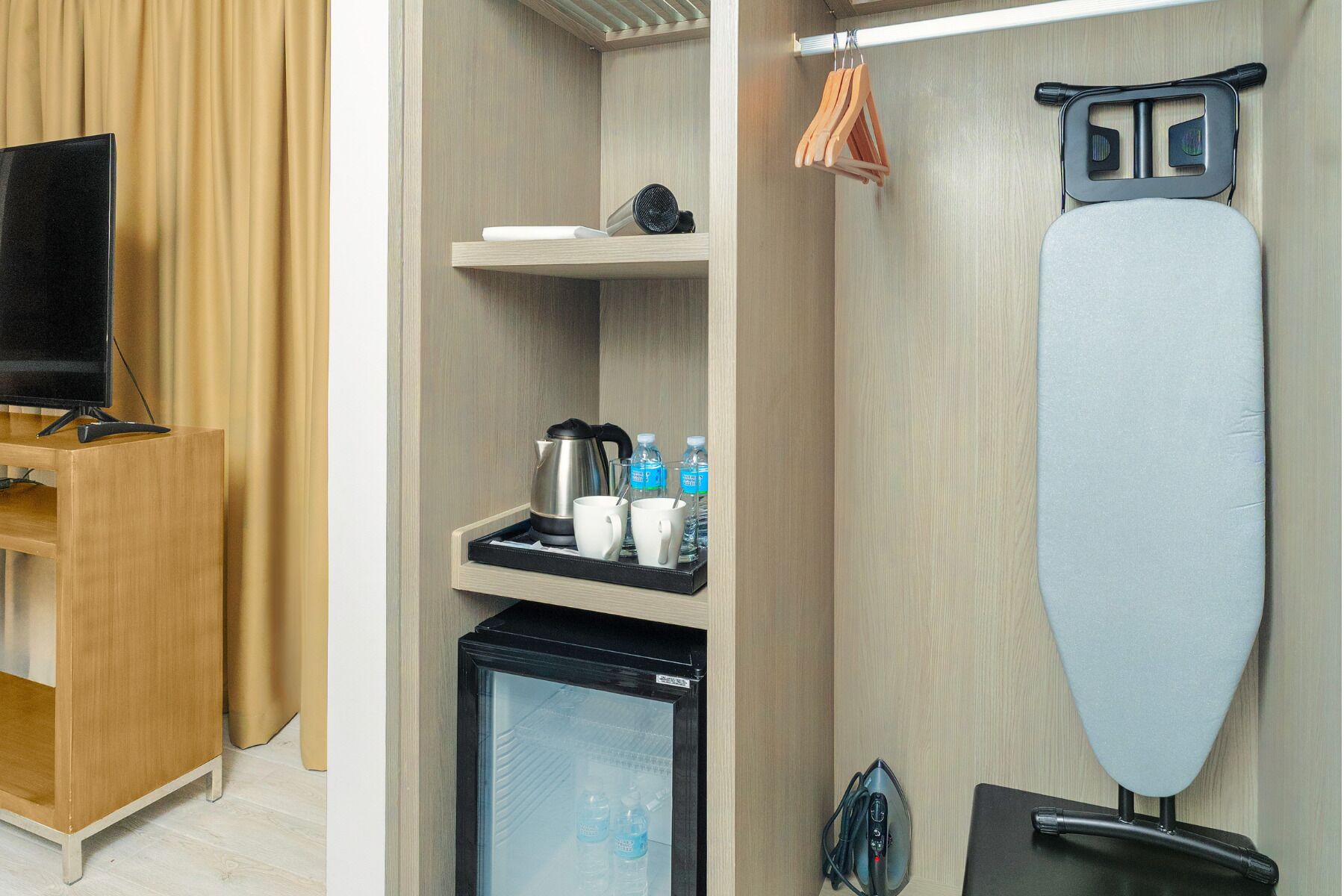 A compact hotel room with a flat-screen TV, a mini-fridge, a coffee maker, and an ironing board.