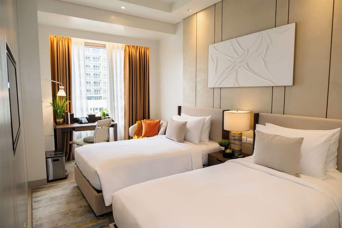 A hotel room with two beds, a desk, a chair, a lamp, and a large abstract painting on the wall.