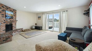 Ein Wohnzimmer mit Kamin aus Backsteinen, einem grauen Sofa, einem gemusterten Teppich und einem großen Fenster mit Vorhängen.