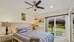Ein modernes Schlafzimmer mit einem großen Bett, einem Deckenventilator, einem Nachttisch und einem Beistelltisch, mit Blick auf einen Garten durch eine Schiebetür.