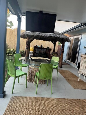 Une terrasse extérieure avec une table ronde en verre et des chaises vertes, une structure dotée d’un toit de chaume et une vue sur une clôture en bambou.