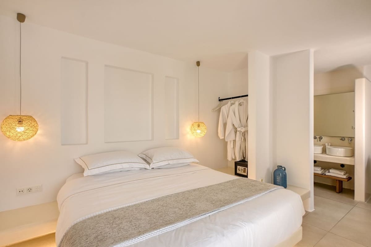 Une chambre à coucher avec un grand lit, deux lampes fixées au mur, un porte-manteau et une salle de bain visible à travers une porte ouverte.