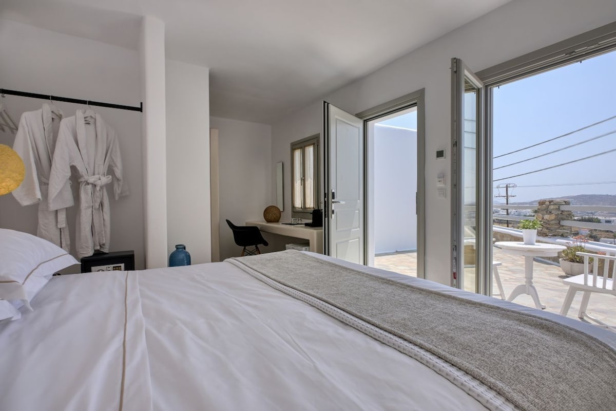 Habitación de hotel con cama, escritorio, silla, baño con espejo y balcón con vistas.