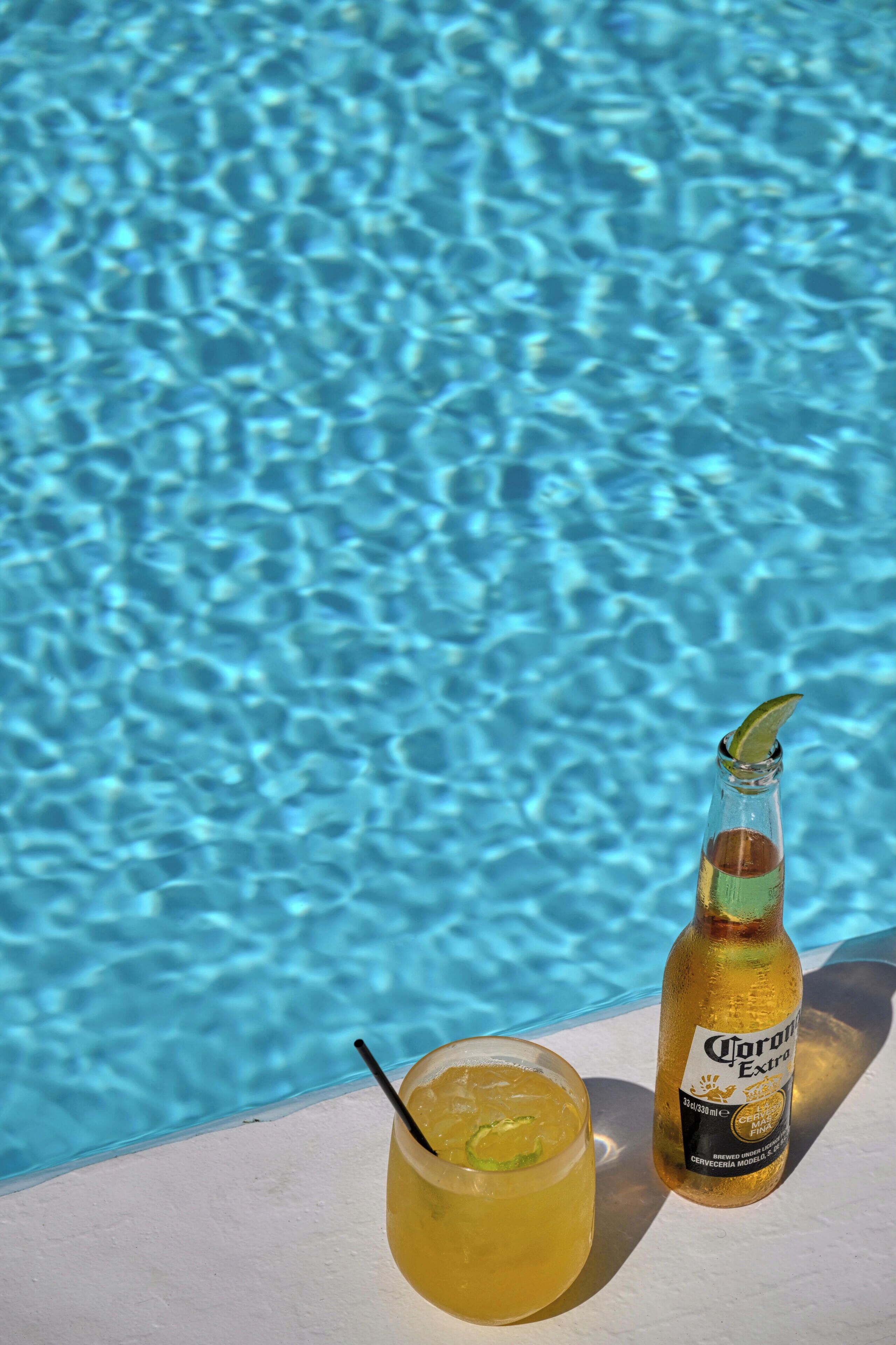 A glass of a yellow beverage with a lime wedge and a bottle of Corona Extra beer by a poolside.