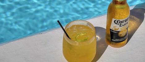 A glass of a yellow beverage with a lime wedge and a bottle of Corona Extra beer by a poolside.