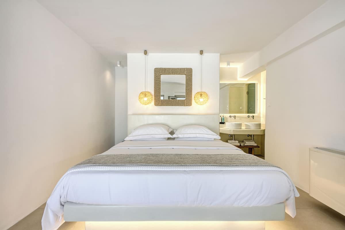 Une chambre d’hôtel moderne avec un grand lit, une table de chevet avec une lampe et une salle de bain équipée d’un lavabo et d’un miroir.