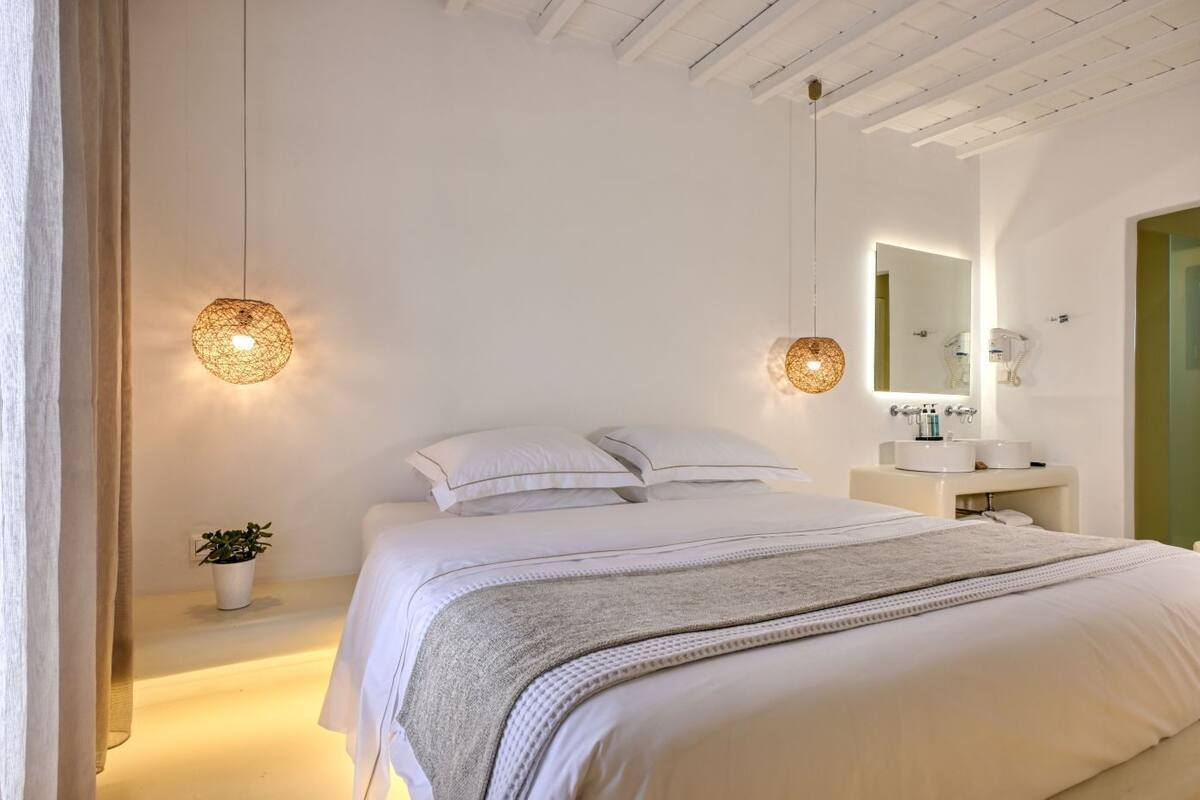Une chambre à coucher avec un lit, une table de chevet agrémentée d’une plante, des luminaires suspendus et une salle de bain avec un miroir et un lavabo.