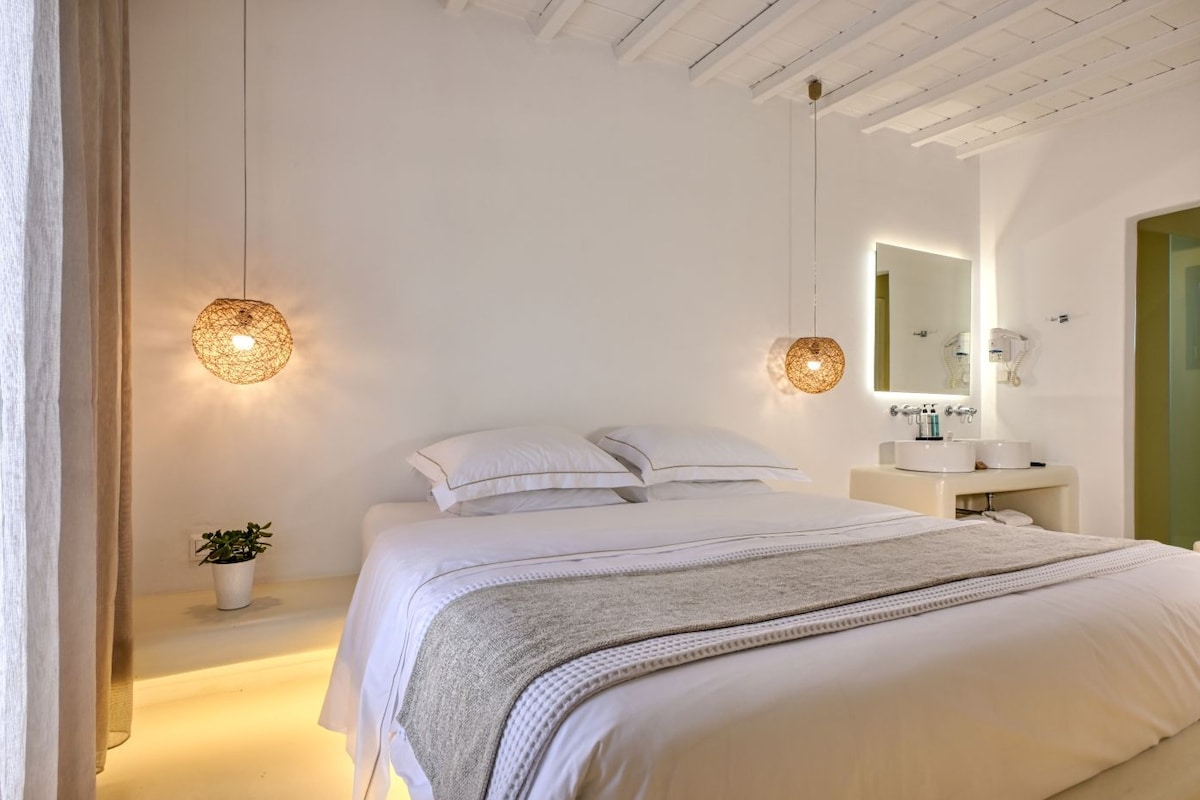 Un dormitorio con cama, mesita de noche con una planta, lámparas colgantes y un baño con espejo y lavabo.