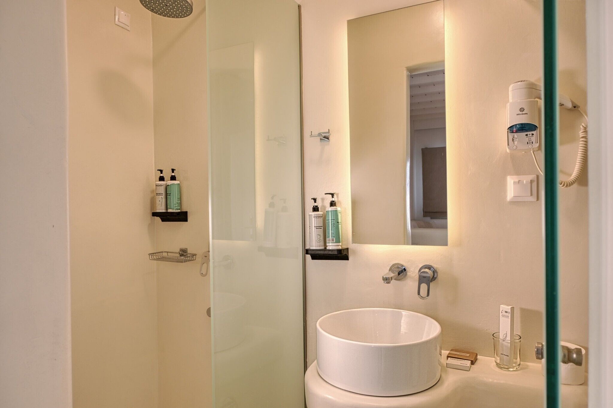 A bathroom with a glass shower enclosure, a white sink, and a mirror. There are toiletries on a shelf and a hairdryer on the wall.
