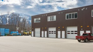 A large industrial building with multiple garage doors, a red pickup truck parked in front, and a mountainous background. - Revy To Go - Unique 2-Bedroom Condo (Revelstoke)
