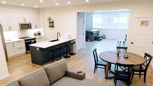 A modern kitchen with white cabinets, a central island with bar stools, a dining area with a round table and chairs, and a view into a living space with a sofa and a TV. - Revy To Go - Unique 2-Bedroom Condo (Revelstoke)